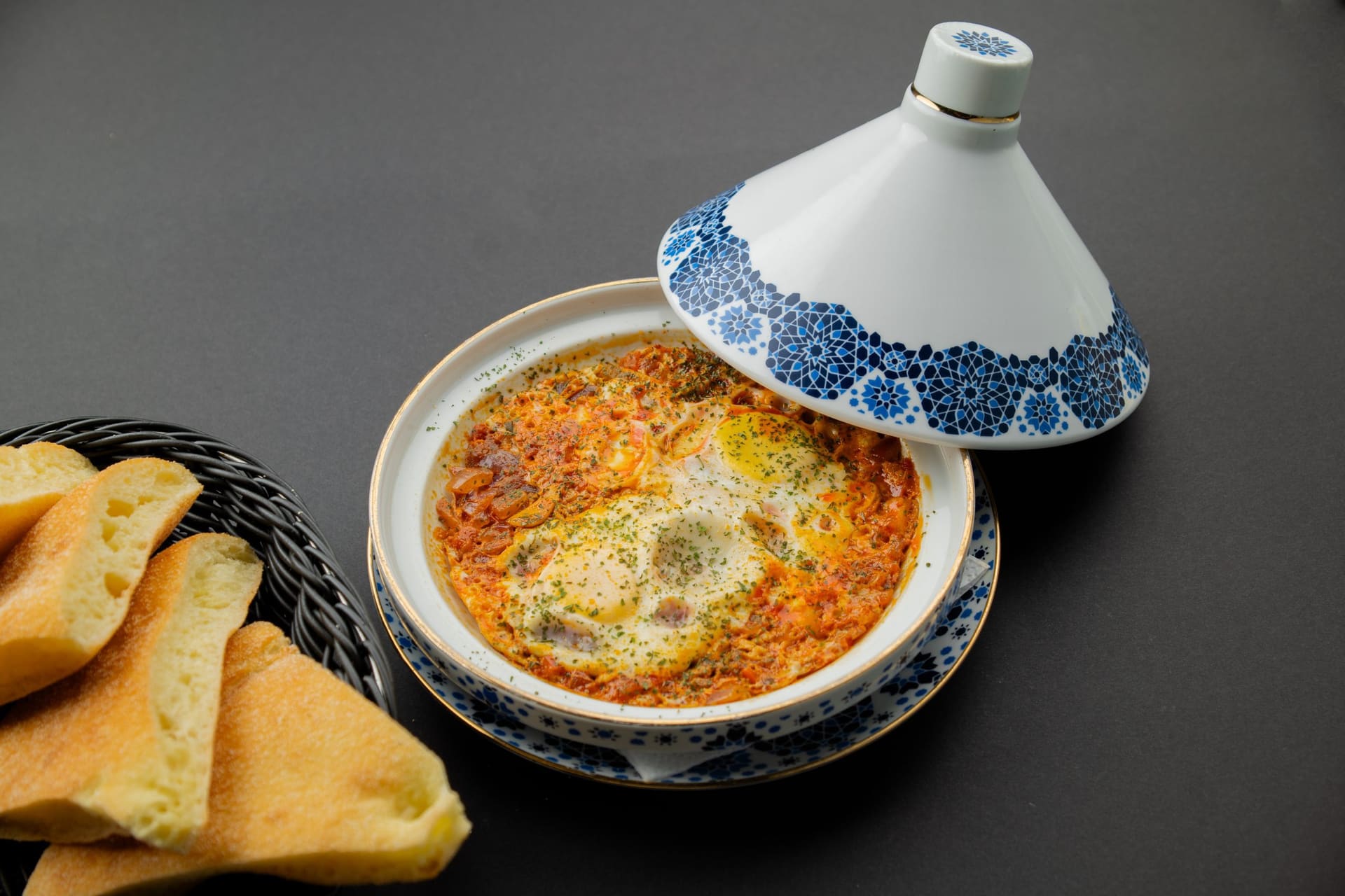 Tajine met eieren in tomatensaus onder een blauw met witte deksel, naast een mand Marokkaans brood